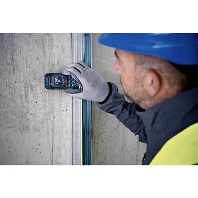 A worker wearing a blue hard hat and gloves is measuring the wall thickness of a concrete wall using a digital measuring device.