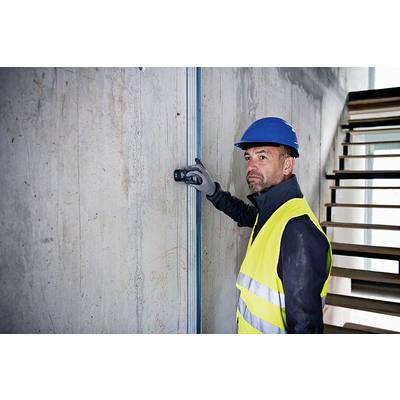 A construction worker in a high-visibility vest and hard hat is measuring a concrete wall in an unfinished building. A staircase is visible in the background.
