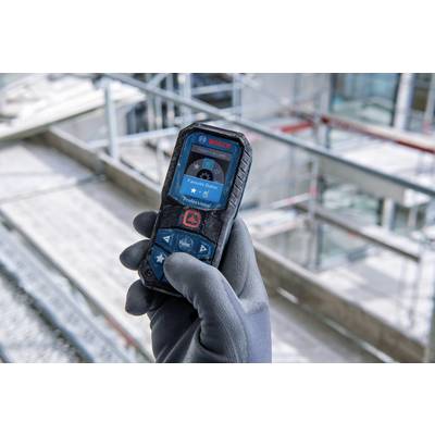 A hand in a grey glove holds a Bosch laser distance measuring device in front of a construction site backdrop. The device displays 'Alignment'.