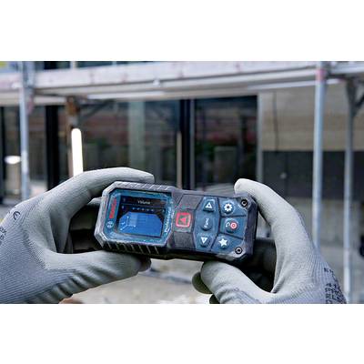 A person is holding a digital distance metre with a scaffolding in the background. The display shows a diagram for volume calculation.