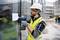 A construction worker wearing a hard hat and high-visibility vest is securing a document to a glass wall using a cordless drill on a building site.