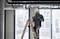 A construction worker wearing a hard hat and safety glasses stands on a ladder in a modern building and drills into the ceiling.