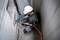 A worker in protective clothing and a hard hat is securing an orange cable to a concrete wall using an electric tool.