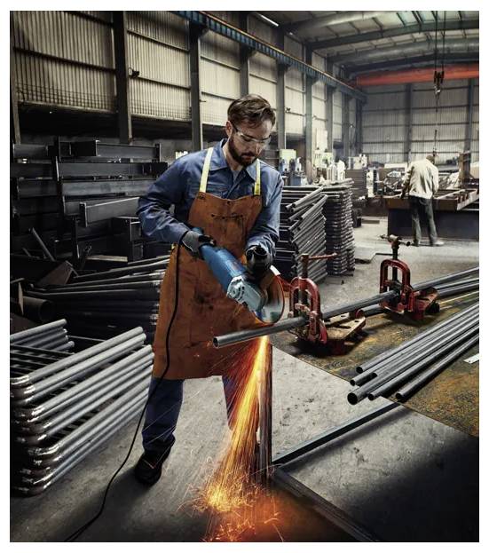 A worker in a workshop uses a power tool to cut metal, with sparks flying. Stacks of metal pipes are visible in the background.