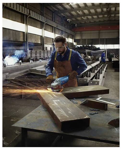 A worker in a workshop wearing safety gear grinds a large metal beam, creating bright sparks. The background shows metal structures and machinery.