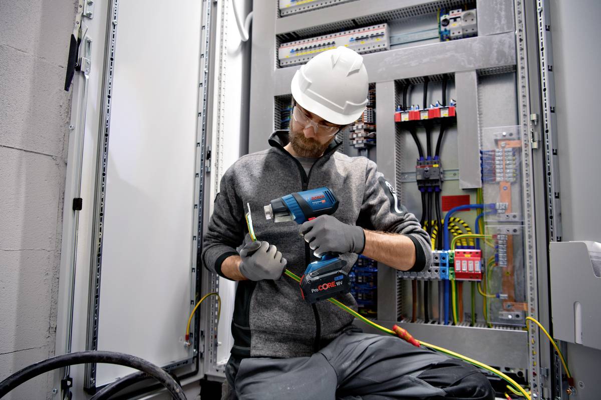 An electrician in work attire and safety helmet is checking cable connections in an open electrical cabinet with various cables and circuits.