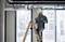 A construction worker is standing on a ladder and using a cordless drill to install cables on the ceiling of an unfinished building.