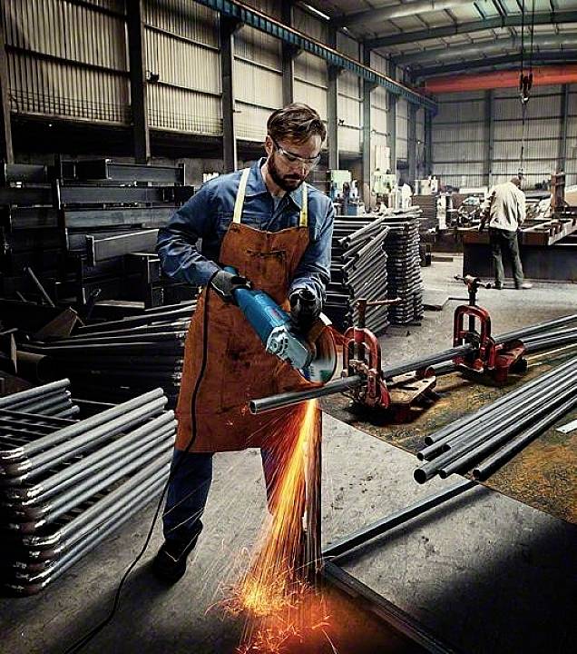 A man in protective clothing is grinding metal in a factory hall, sparks flying from the machine. Background of metal pipes.