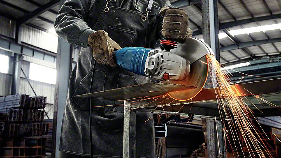A person wearing an apron cuts a metal plate with an angle grinder; sparks are flying to the side. Workshop environment in the background.