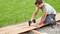 A man is building a wooden decking on a gravel surface using a cordless drill. Grass can be seen in the background.
