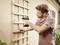 A man is attaching a wooden trellis to a house wall using a cordless drill. He is wearing safety glasses and a shirt.