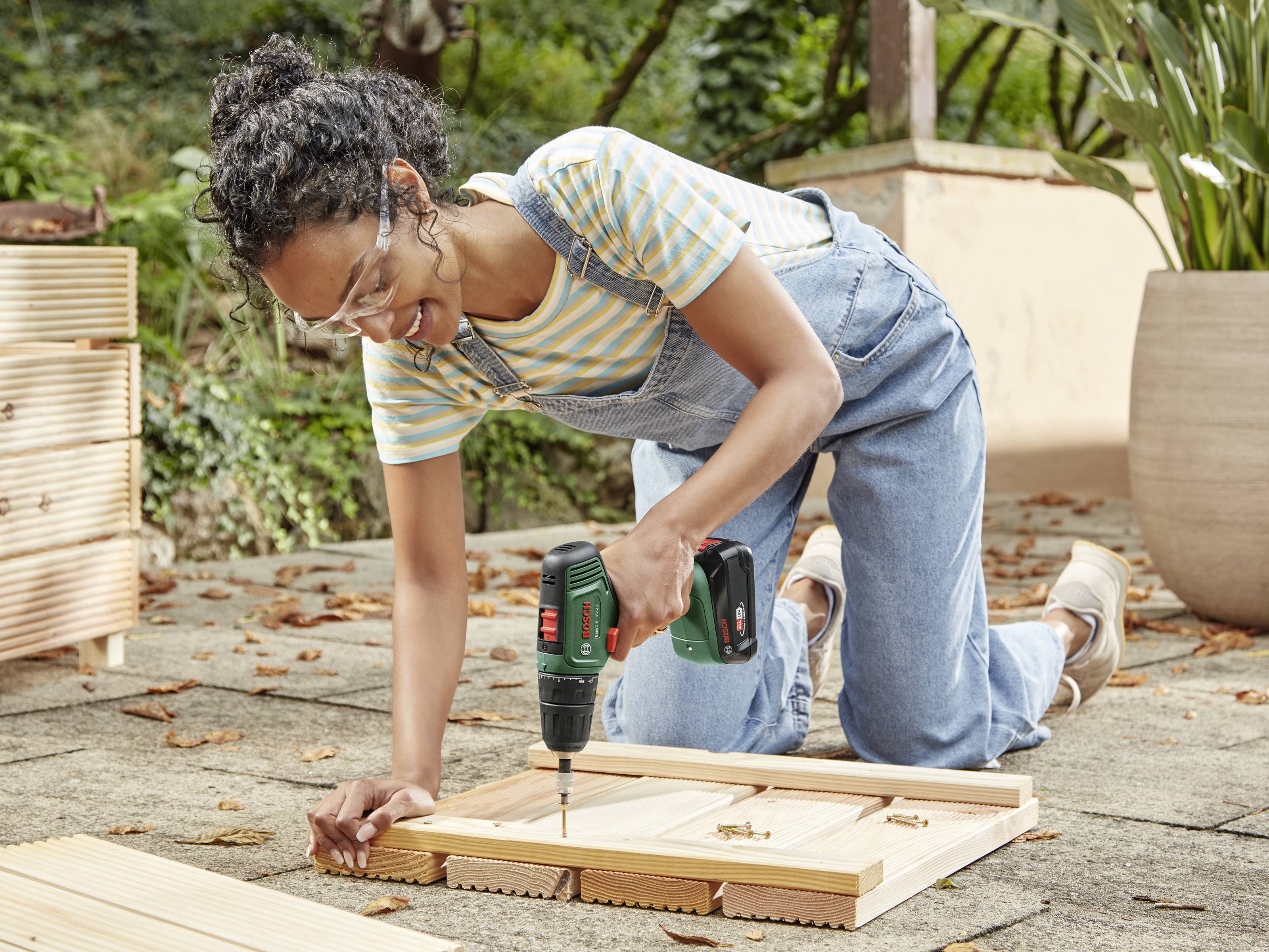 A person is kneeling on the ground and drilling a piece of wood together using a drill. They are wearing safety glasses and dungarees.