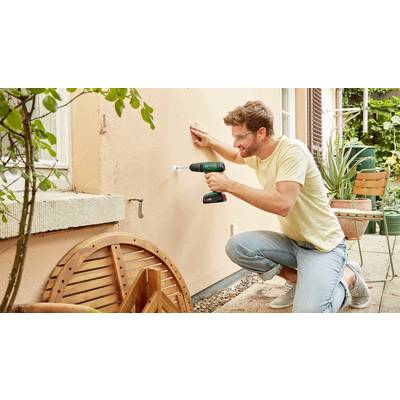 A man is kneeling outside and drilling a hole in a house wall. Next to him are plants and garden furniture.