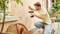 A man is kneeling outside and drilling a hole in a house wall. Next to him are plants and garden furniture.