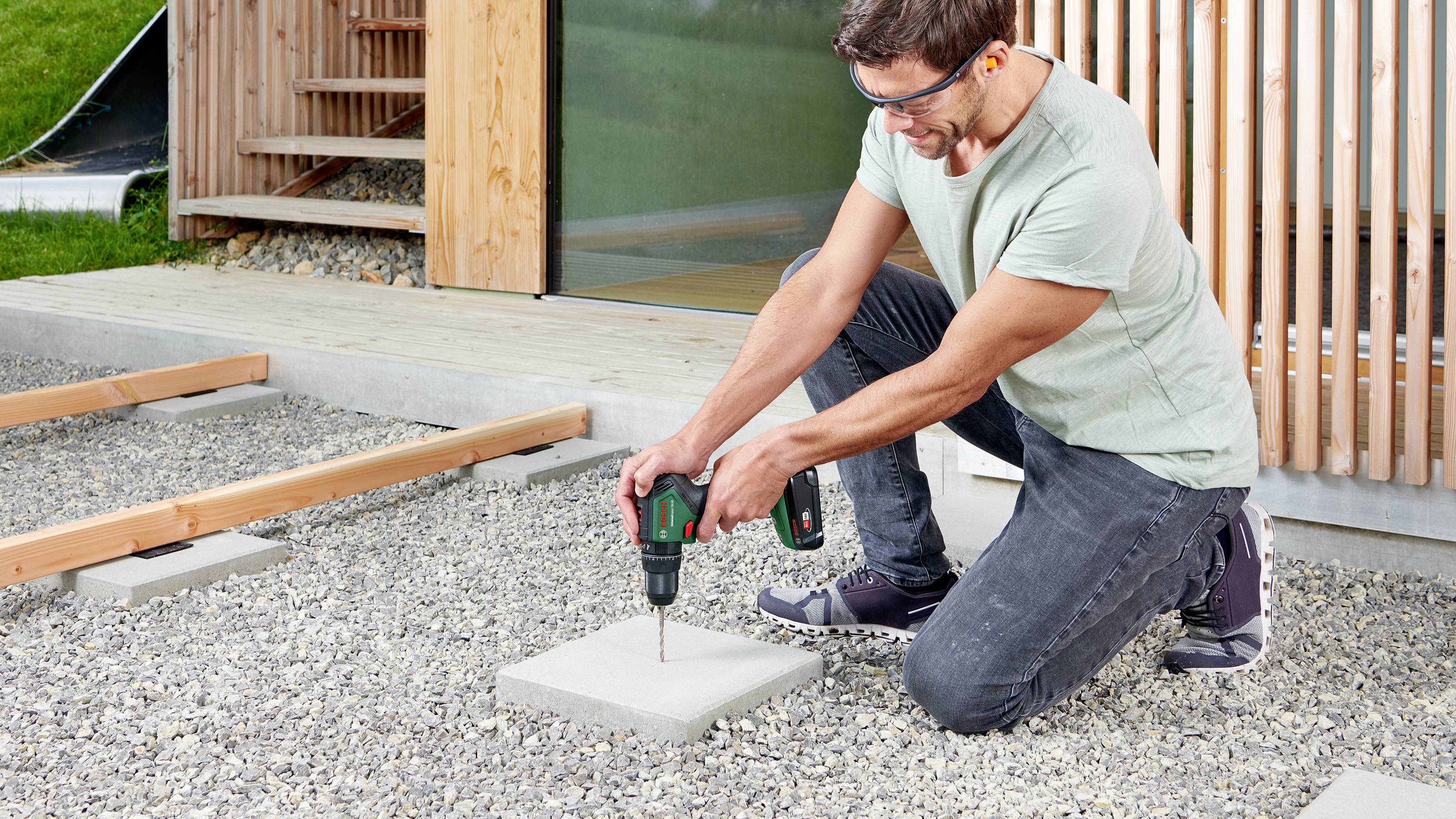 A man kneels outdoors and drills a hole in a concrete slab on a gravel surface, wearing safety glasses.