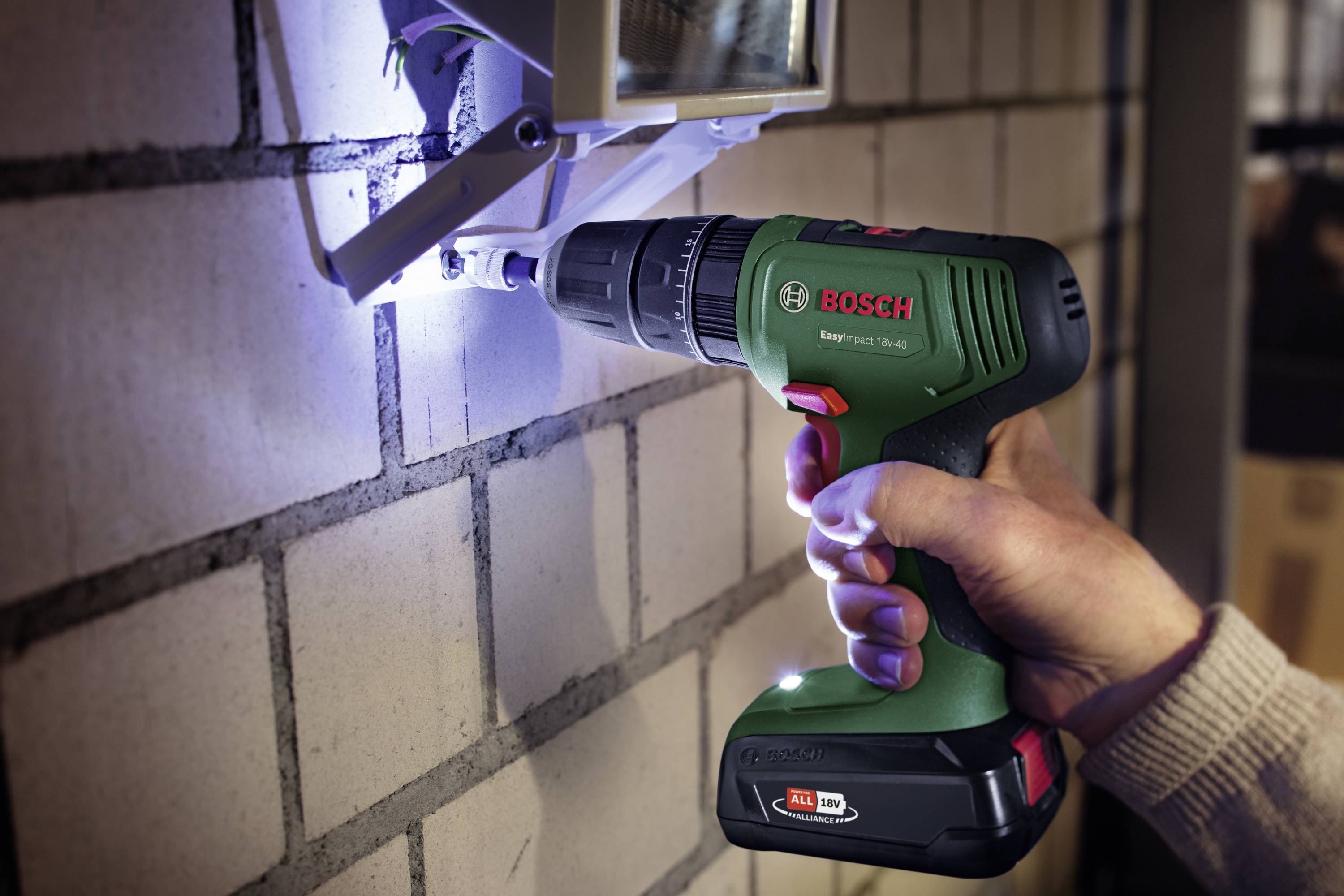 A person is using a Bosch cordless drill to secure a device to a brick wall. The drill illuminates the work surface.