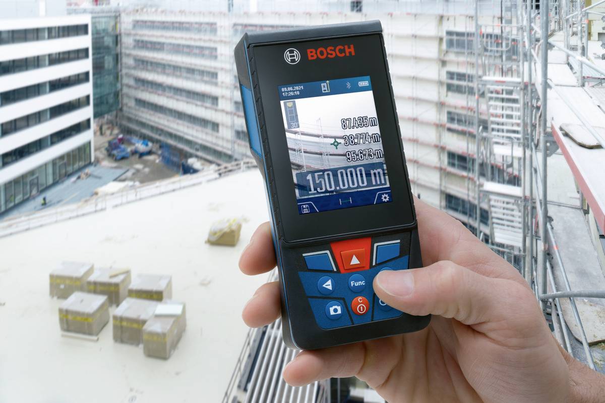 A person is measuring the distance to a building on a construction site using a Bosch laser distance meter. The display shows '150,000 m'.