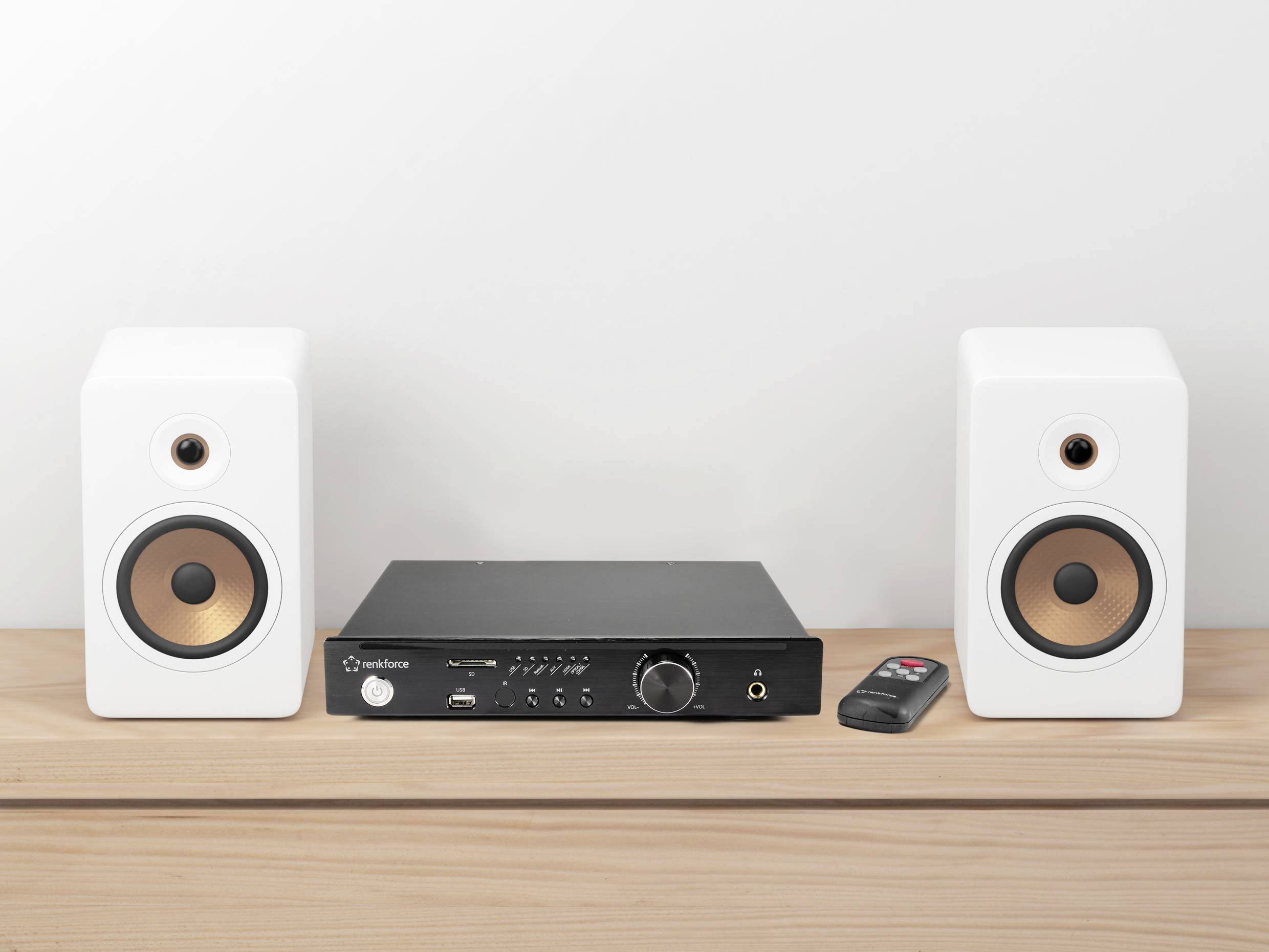 Two white speakers with copper-coloured membranes flank a black amplifier on a wooden shelf. A remote control sits beside them.