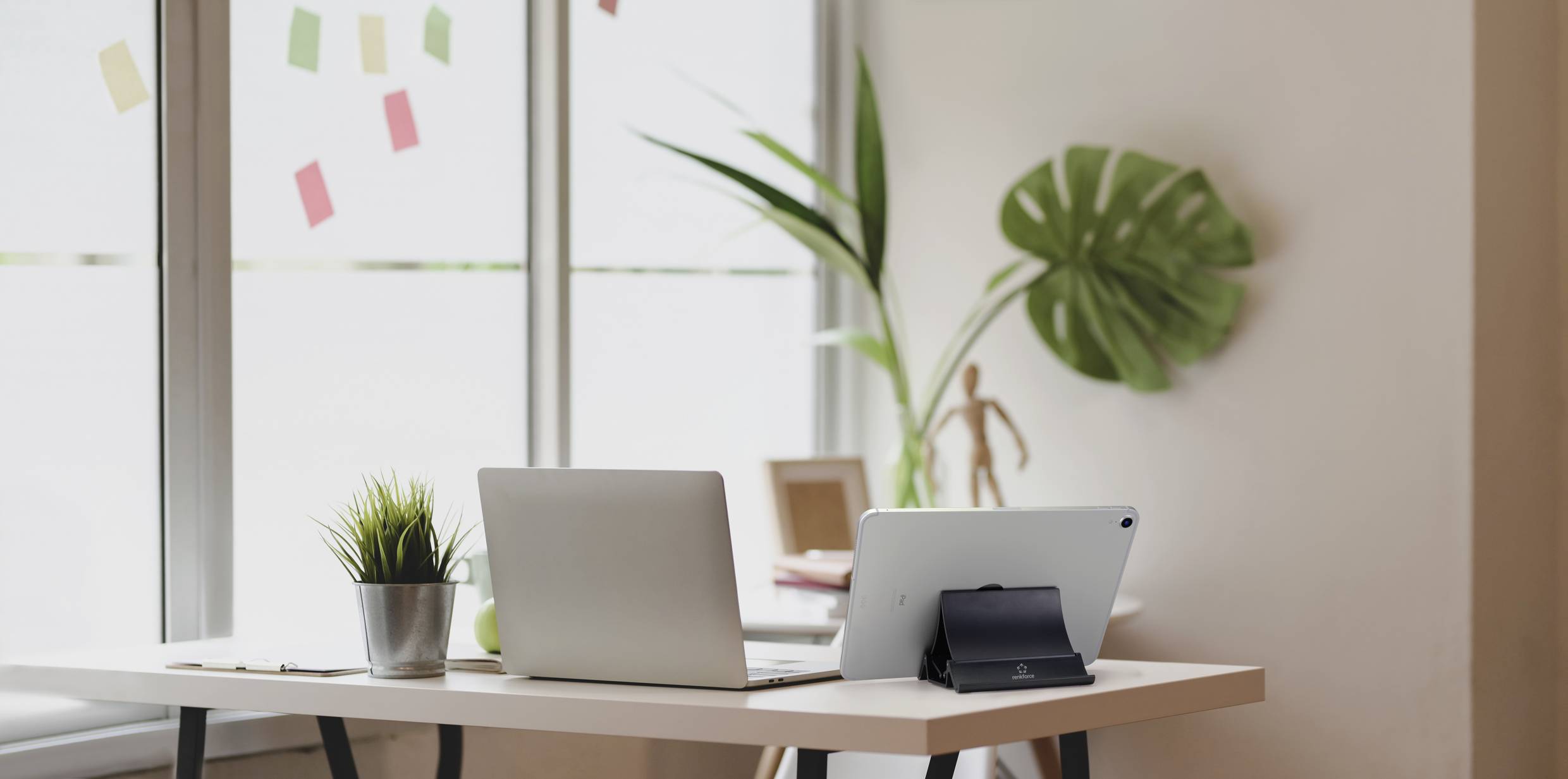 A modern workspace with a laptop and tablet on a white table, surrounded by plants and near a large window with sticky notes.