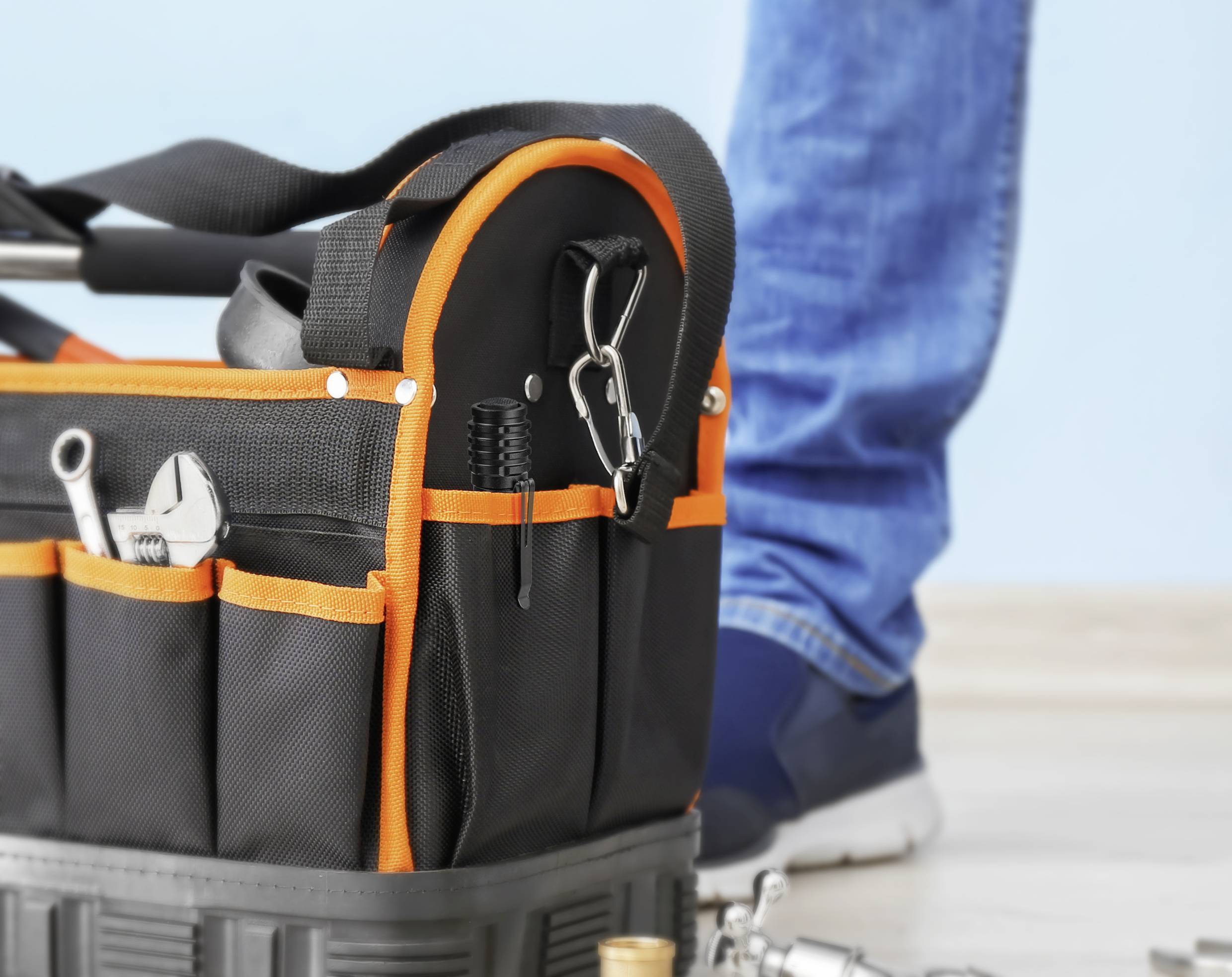 A black tool bag with an orange border sits on the ground. Next to it stands a person wearing blue jeans and trainers.