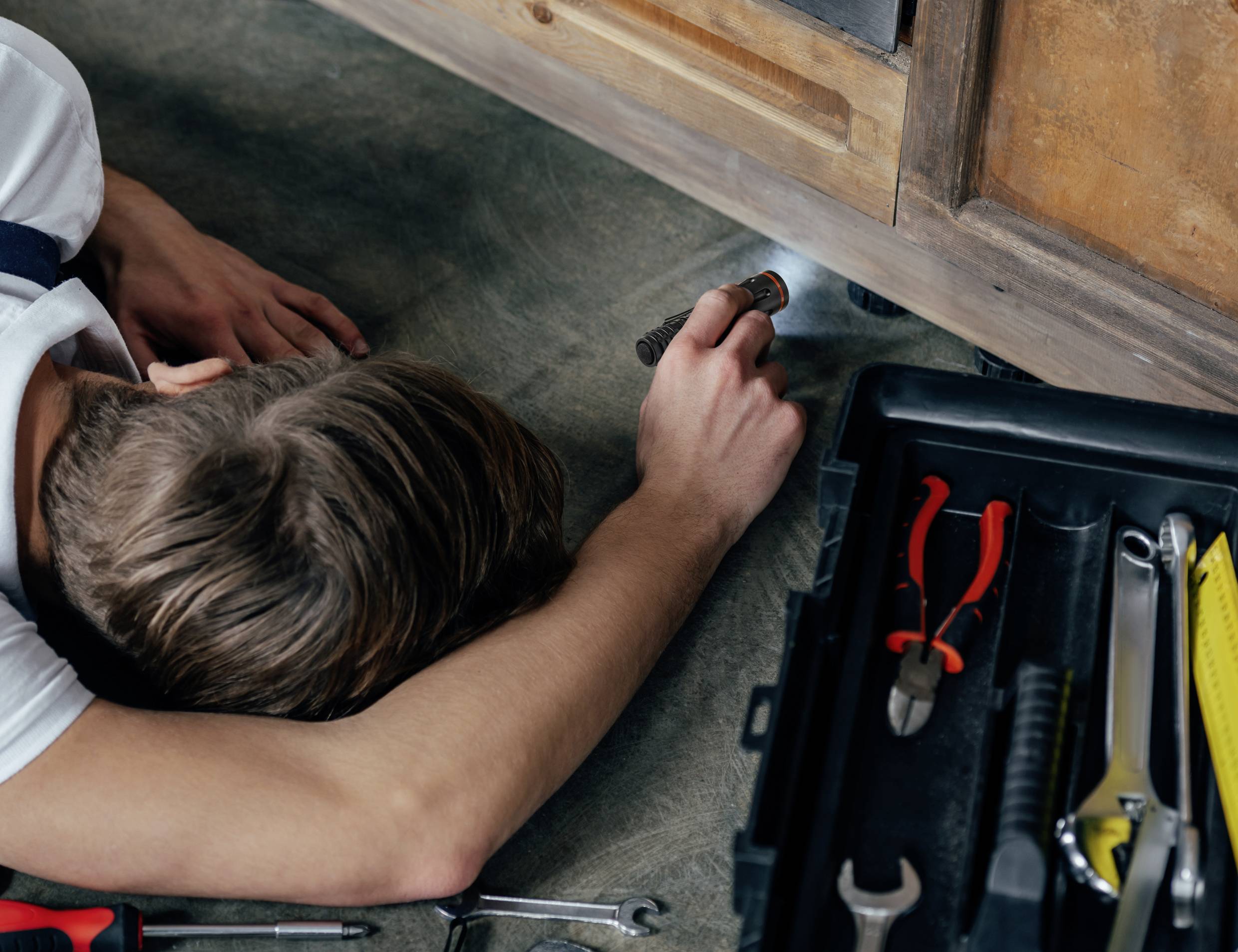 A technician is lying on the floor and shining a torch underneath a piece of furniture. Next to him is an open toolbox.