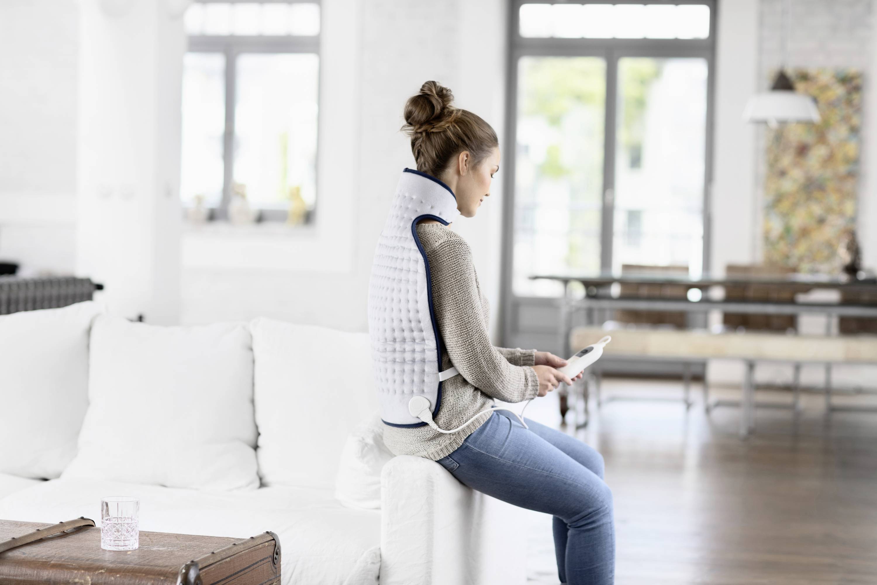 A woman is sitting on a sofa wearing a heated back vest and holding a tablet in a bright living room.