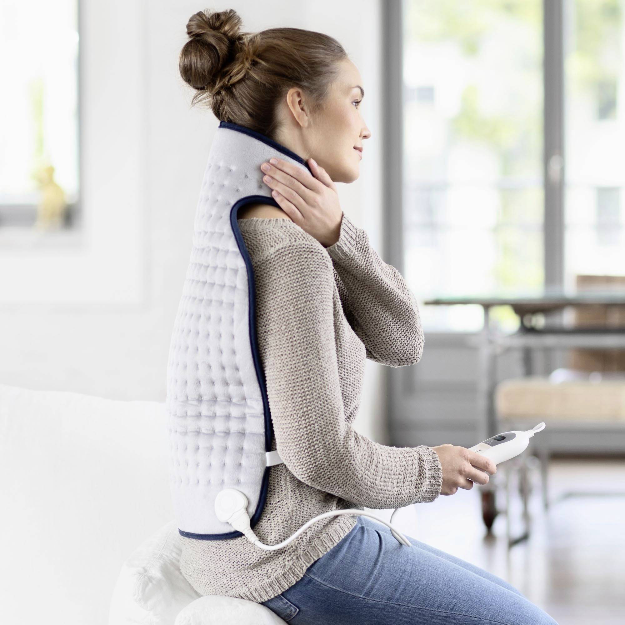 A woman is sitting on a sofa wearing a heated back cushion. She is holding the device's remote control.