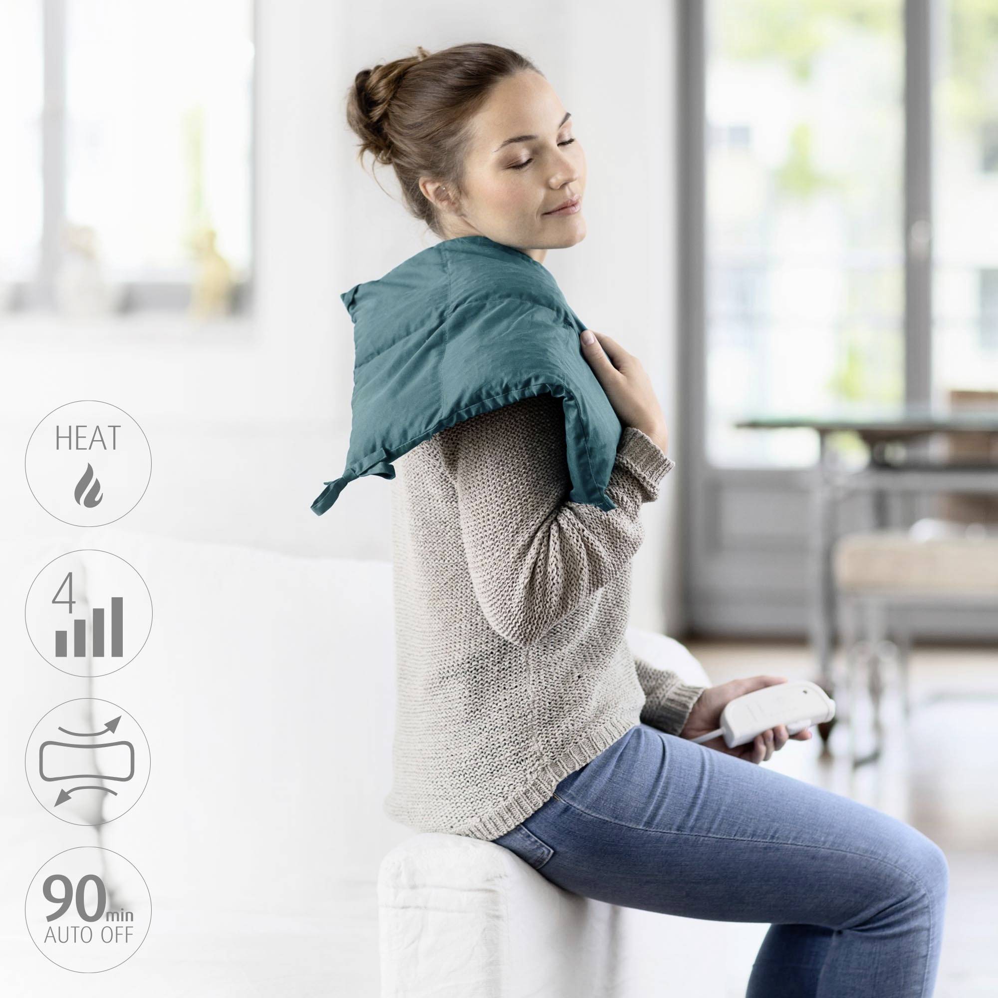 A woman is sitting on a sofa and using a heated neck and shoulder cushion. She is holding a remote control in her hand.