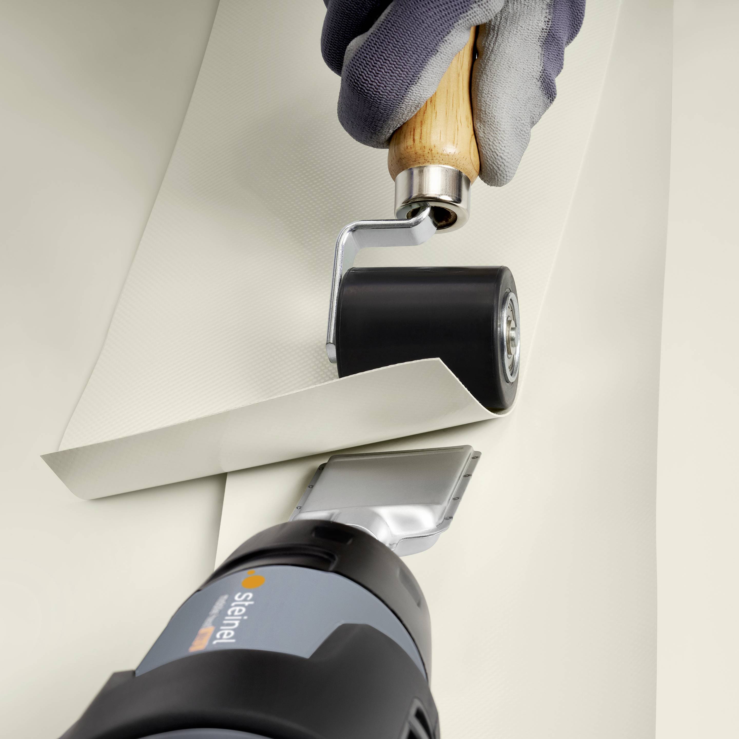 A craftsman is using a roller apparatus to seal an overlap of thick, light-coloured material with a heating tool.