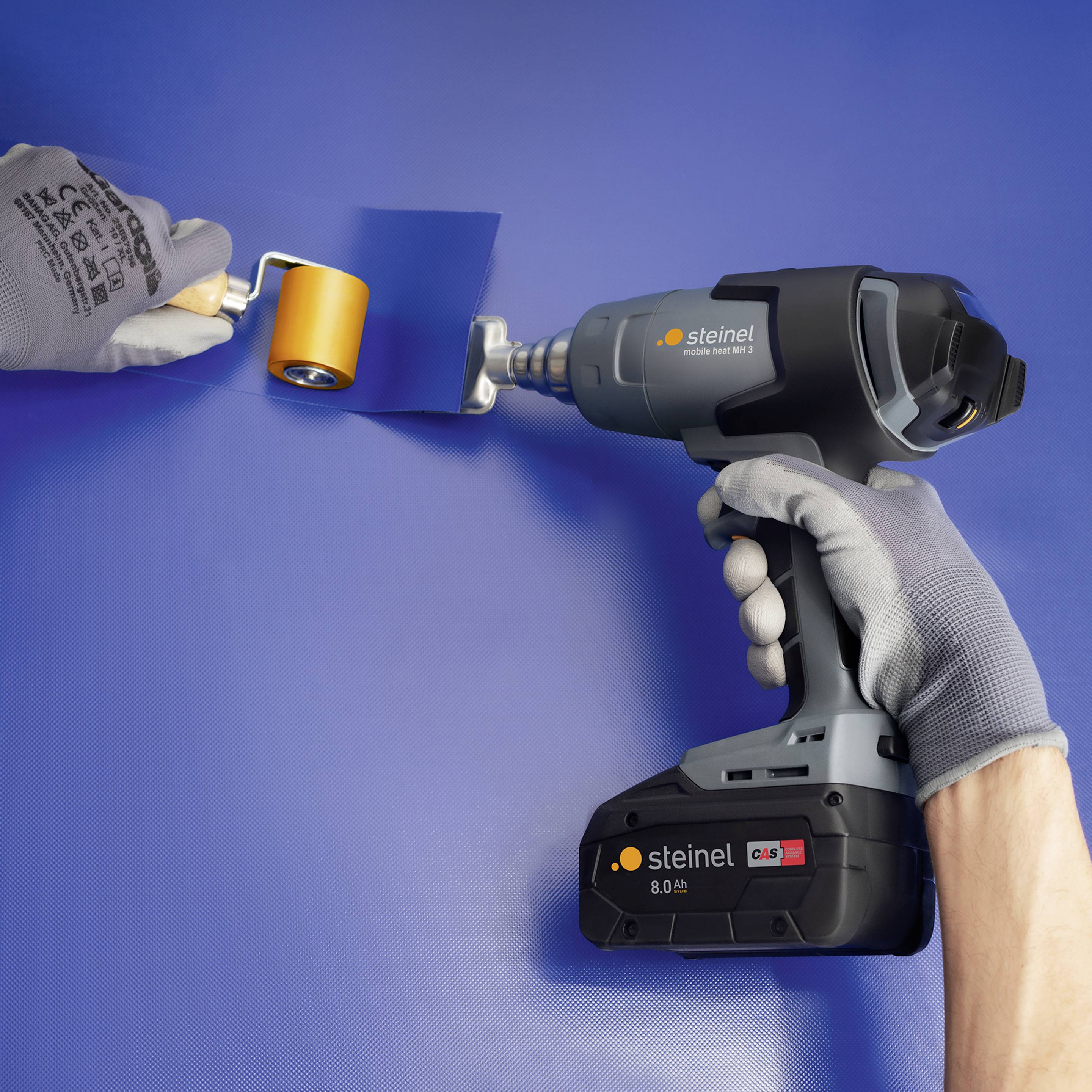 A person is working on a blue surface with a hot air gun and a roller. The devices are black and are from the Steinel brand.