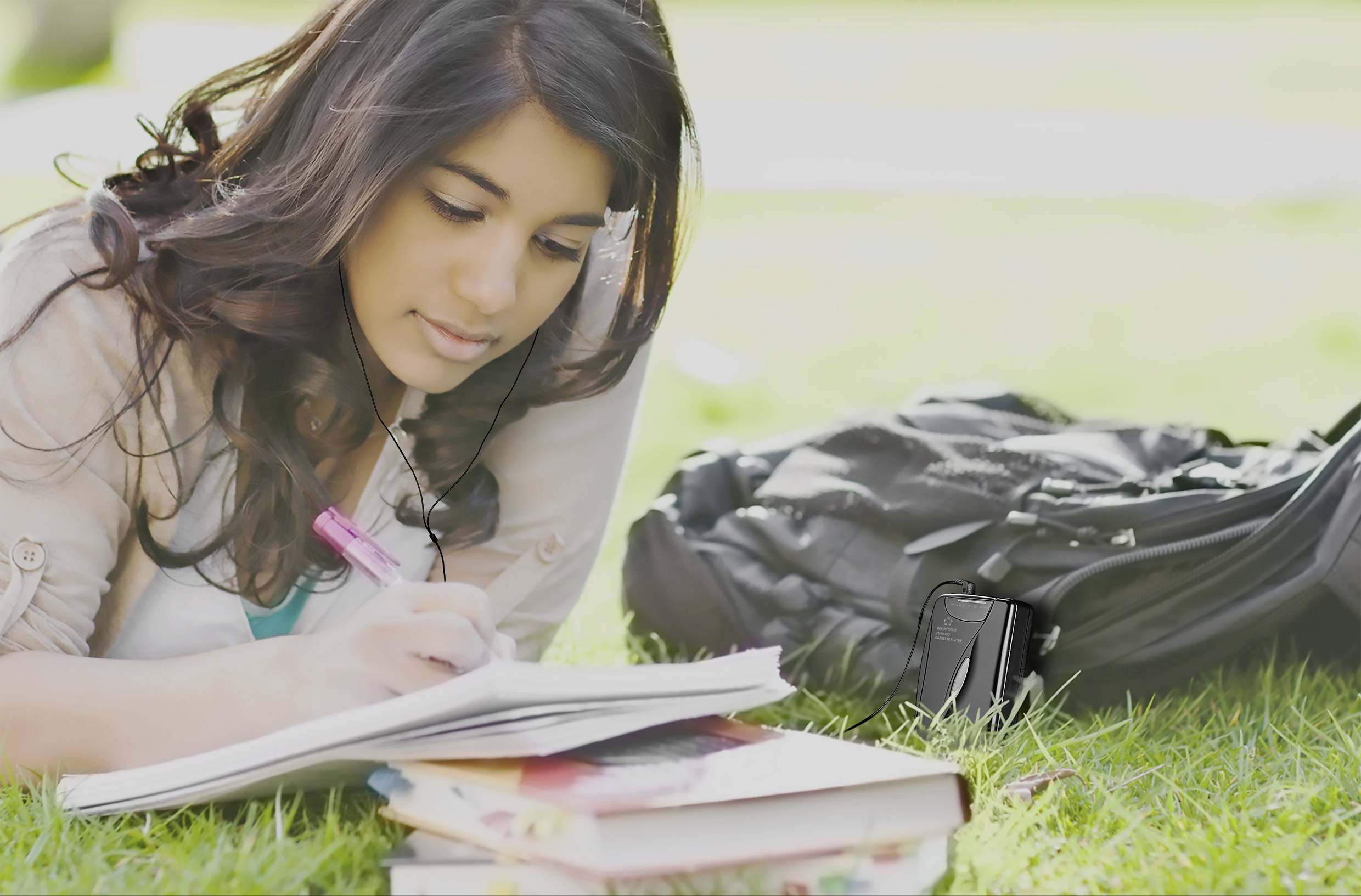 A woman is lying on a meadow and writing in a notebook. Next to her is a black rucksack and several books.