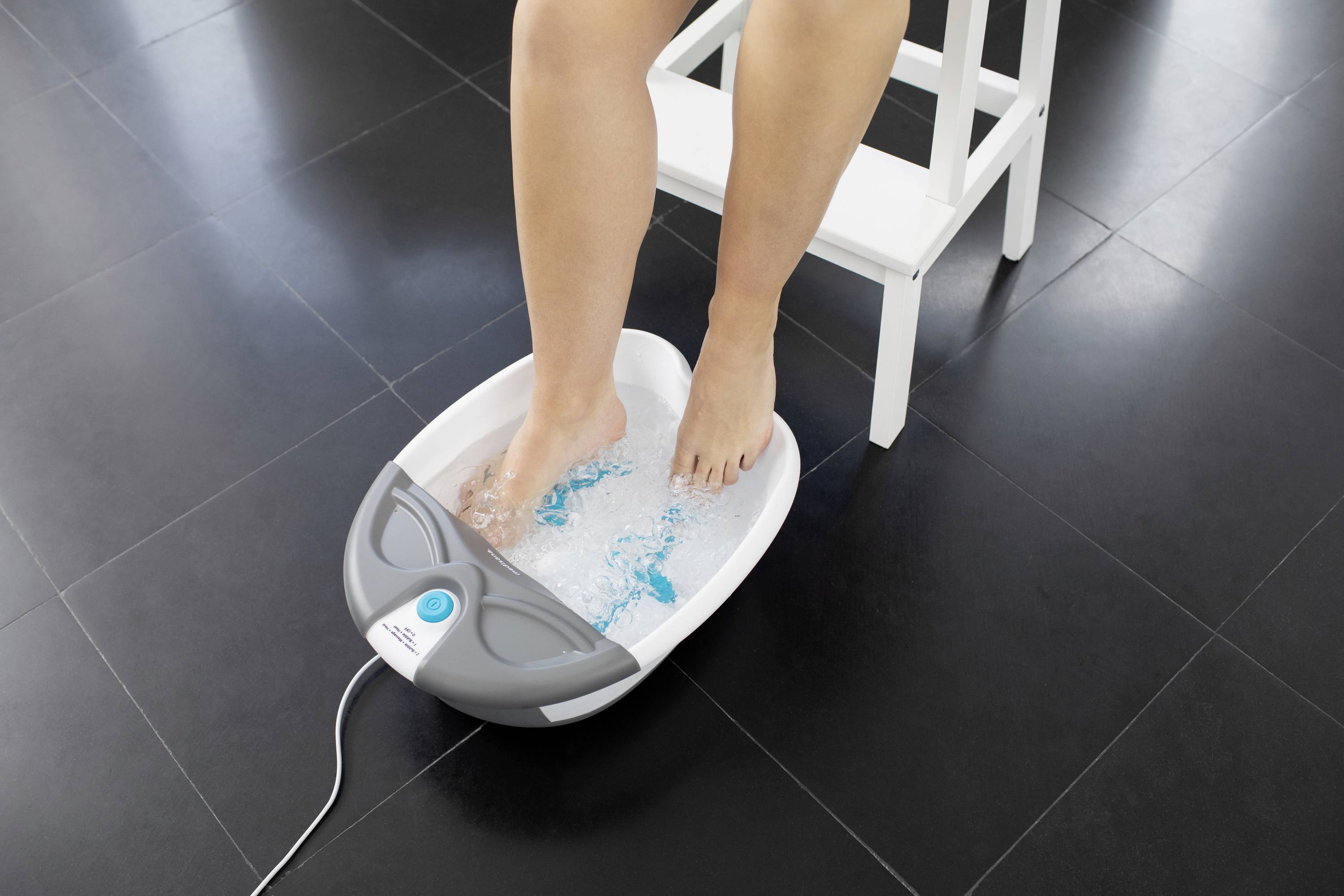 A person relaxes their feet in a bubbling foot bath on a dark grey floor, while sitting on a white chair.
