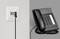 A black cable is connected to a telephone socket, next to a desktop telephone on a grey background.