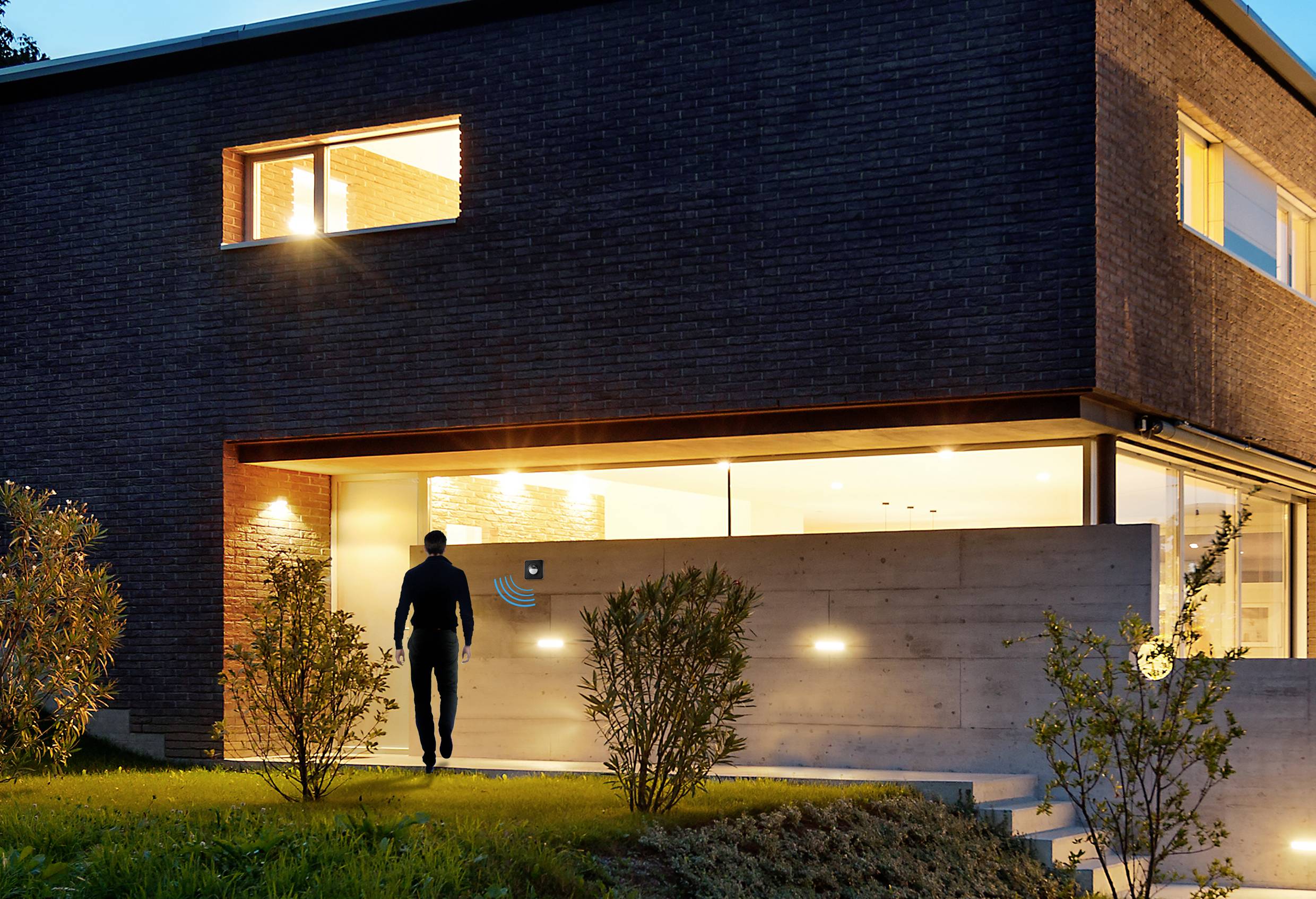Modern brick villa at night with illuminated entrance. A person approaches the door, surrounded by well-maintained shrubs.