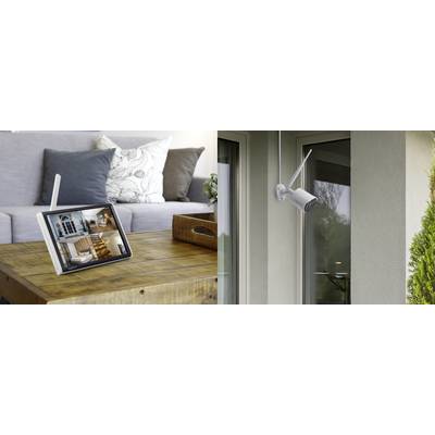A tablet on a wooden table displays camera images of interior spaces. Outside, a security camera is mounted on a house wall.