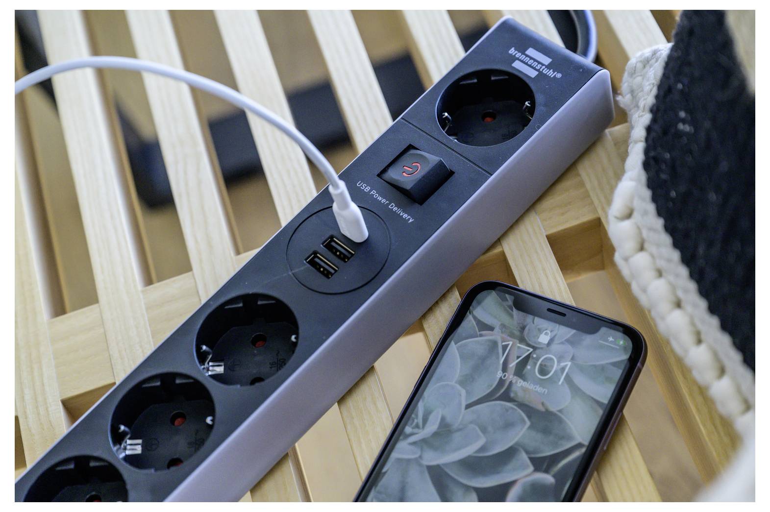 Power strip with a USB charging cable plugged in beside a smartphone on a wooden slatted surface.