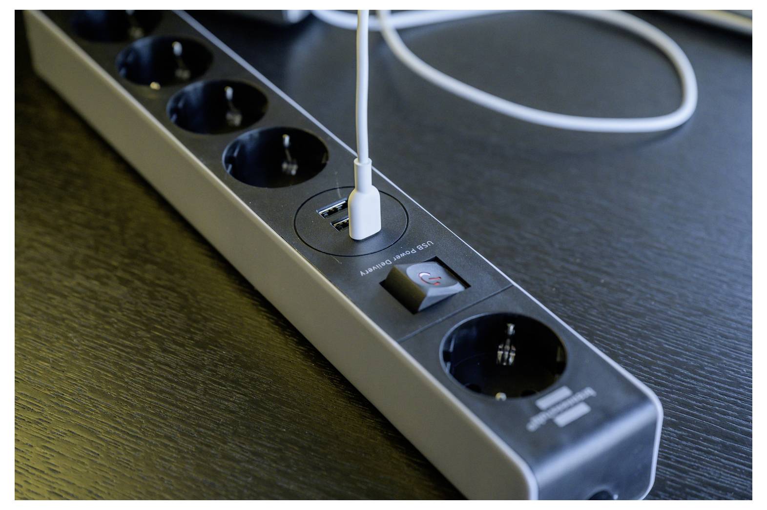 Close-up of a black power strip on a desk, with a USB cable plugged into the built-in charging port and a power switch visible.