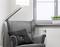 Grey wingback armchair with a black cushion next to a standard lamp in front of a window with white venetian blinds; a radiator is positioned nearby.