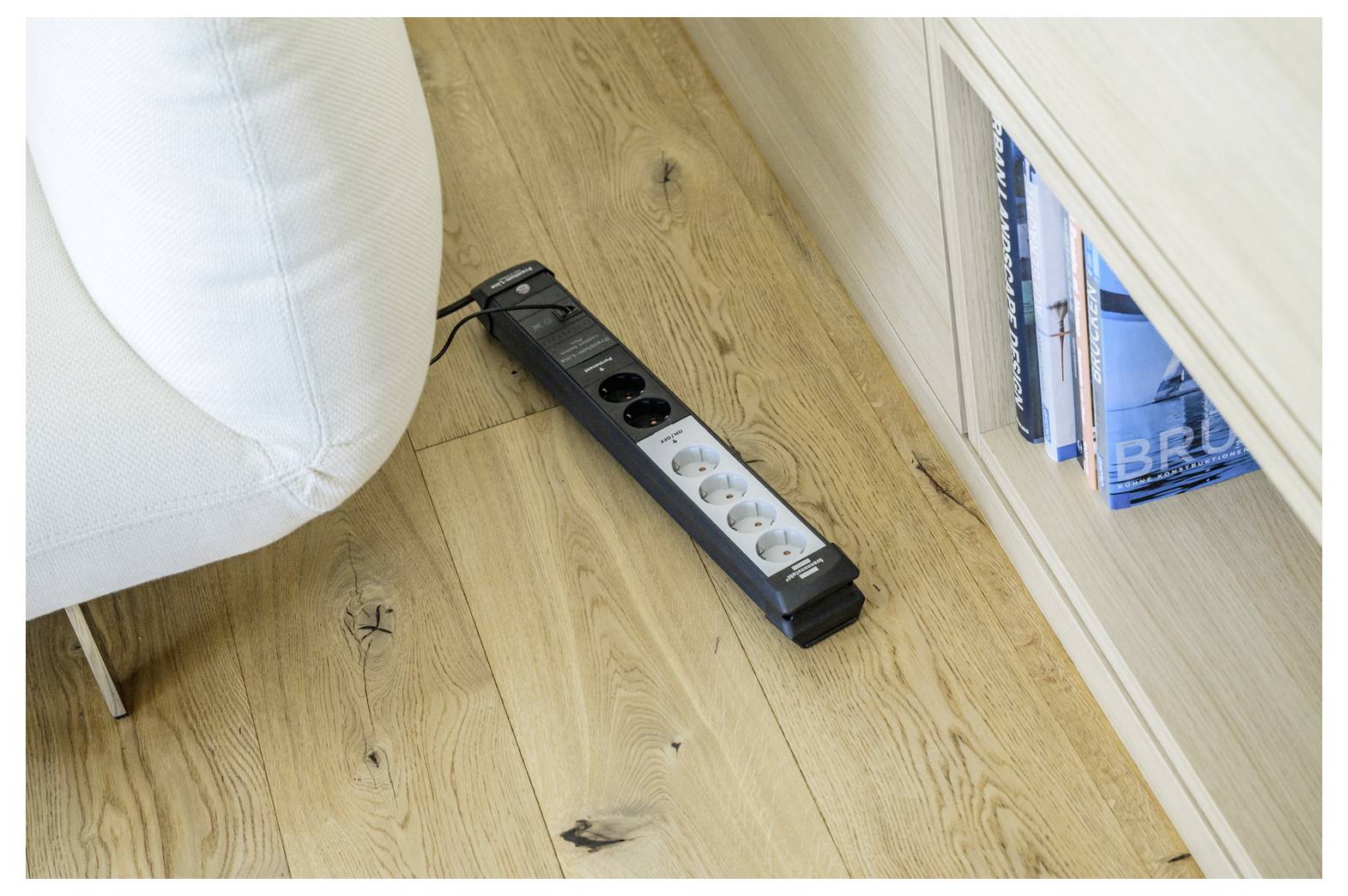 Black power strip with several outlets lying on a wooden floor beside a white sofa and a bookshelf.