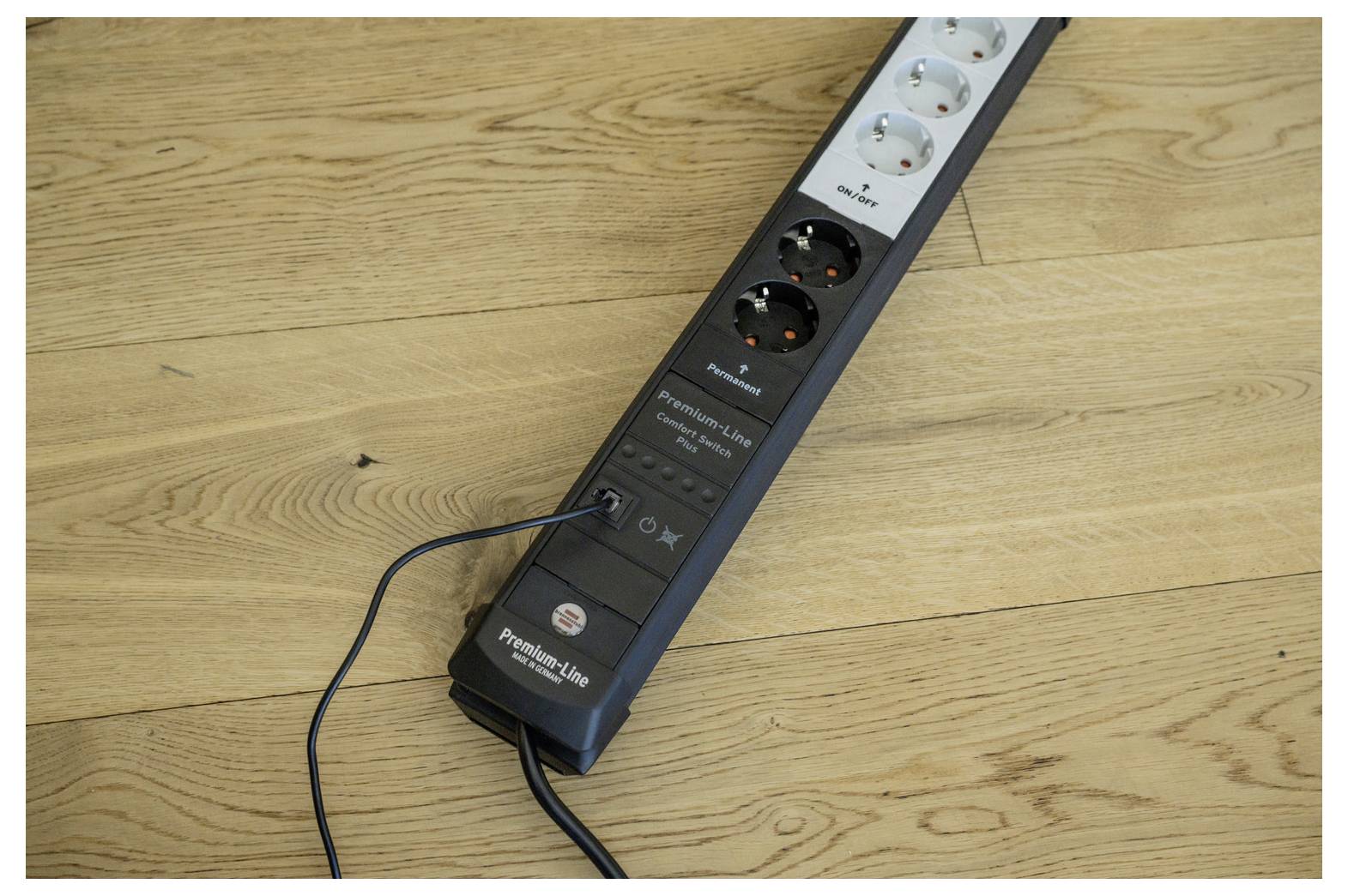 Power strip with several outlets plugged in, lying diagonally on a wooden floor with cords attached.