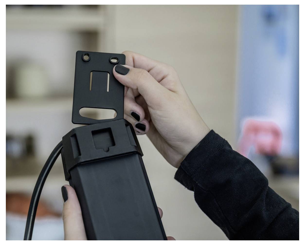 Hand holding a black device bracket above a mounted camera or sensor, showing how the part attaches to the stand.