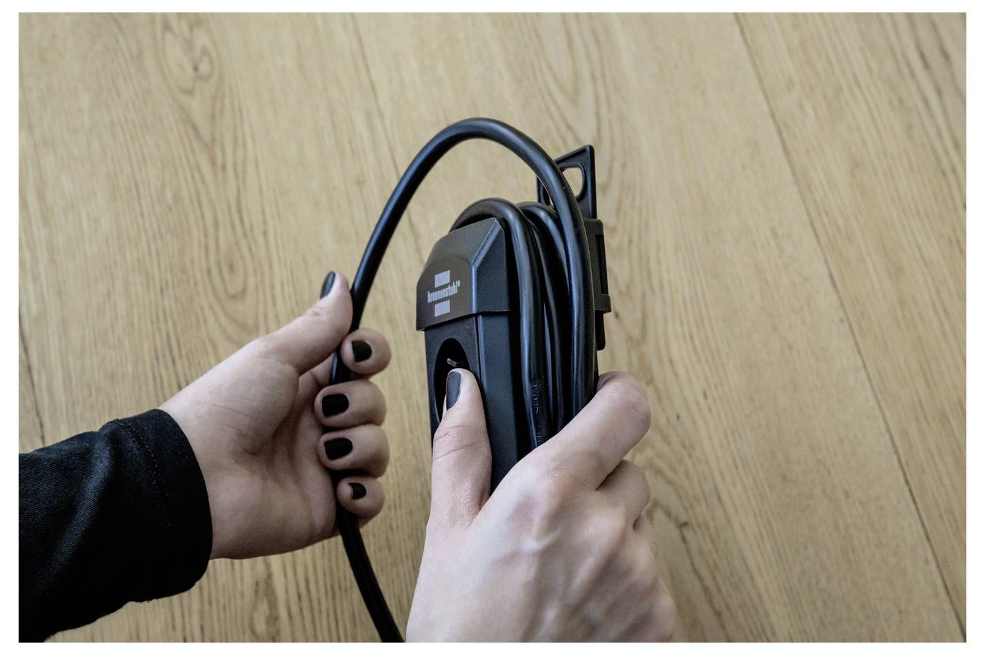 Hands coiling a black cable around a wall-mounted holder on a wooden surface.