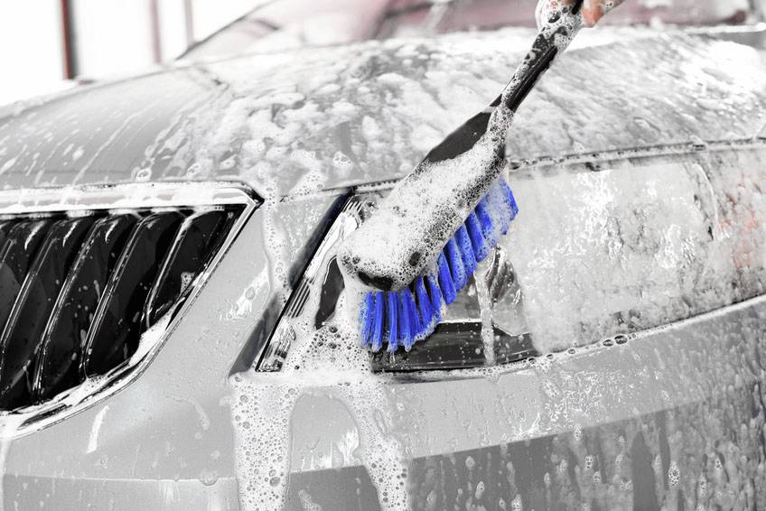 A hand is washing the front of a silver car with a blue brush, which is covered in soapy water.