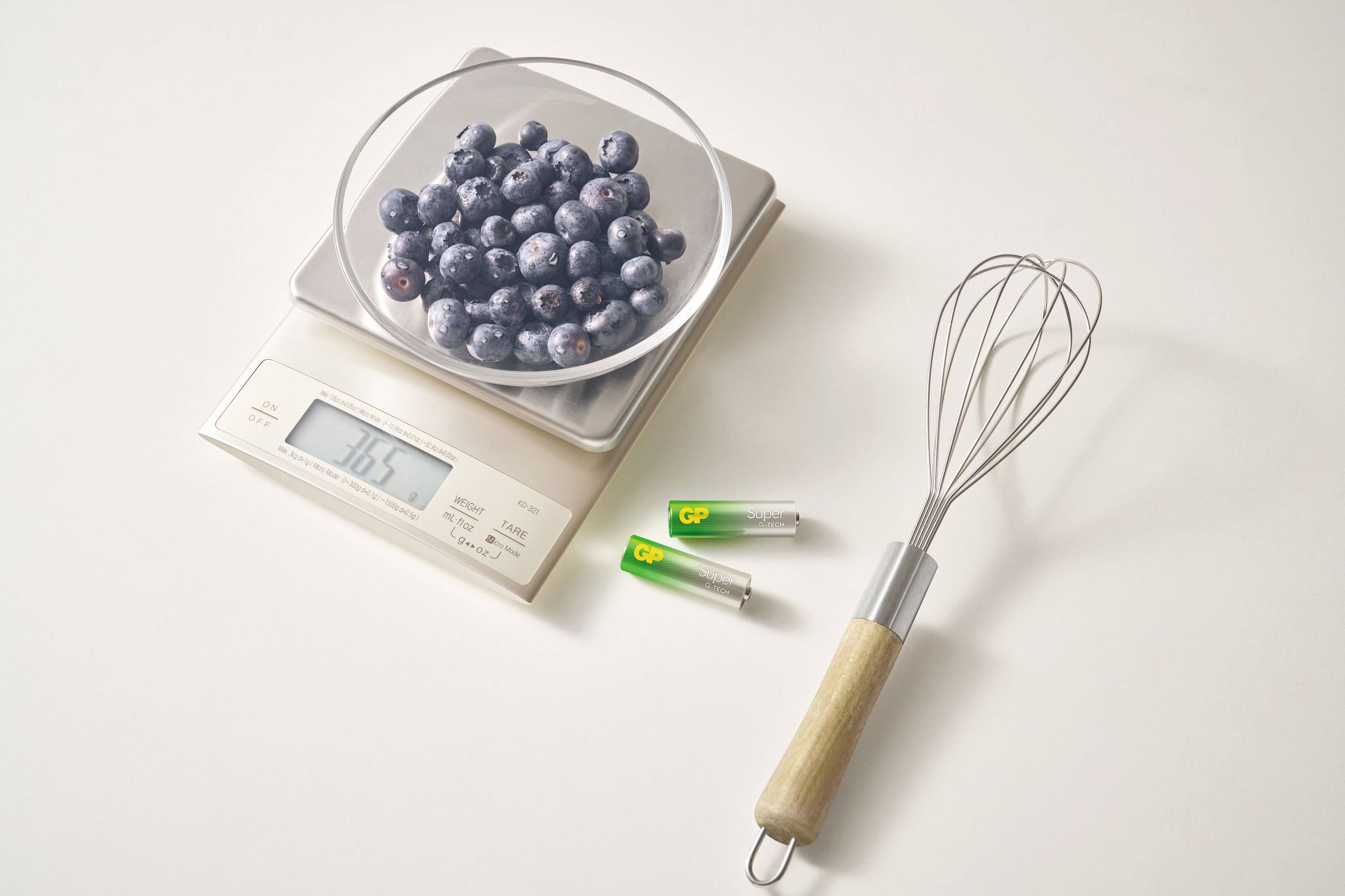 'A bowl of blueberries on a kitchen scale shows 165 grams. Beside it lies a whisk and two empty batteries on a light work surface.'