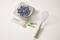 'A bowl of blueberries on a kitchen scale shows 165 grams. Beside it lies a whisk and two empty batteries on a light work surface.'