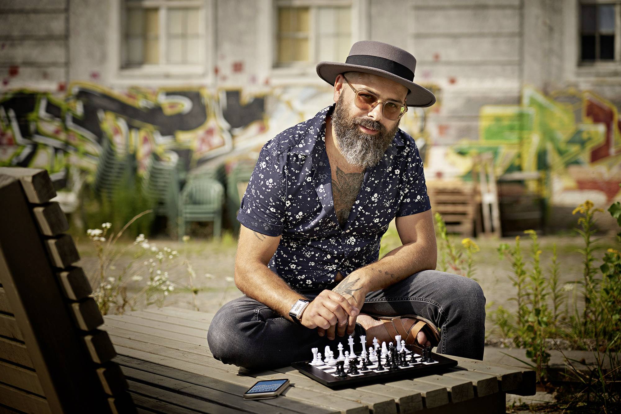 A man wearing a hat sits outdoors on a wooden bench in front of a graffiti wall backdrop, playing chess.