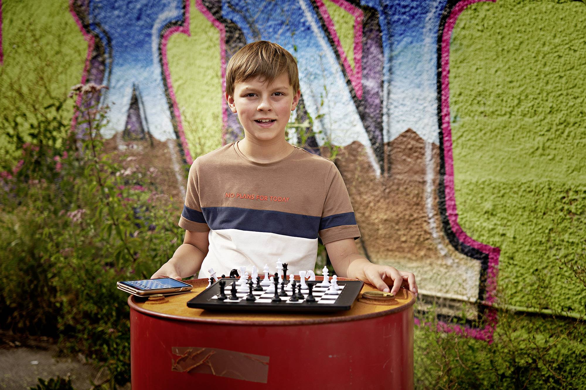 A boy stands outside in front of a colourful wall, holding a chessboard with pieces on a round table, smiling at the camera.