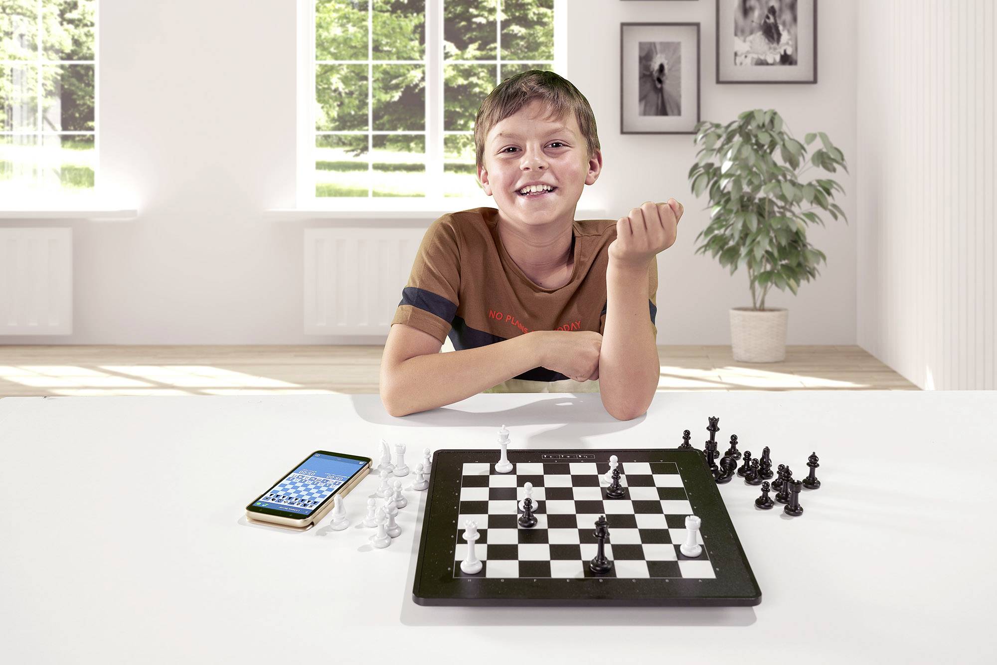 A boy sits at the table, smiling over a chessboard with a smartphone on the table. In the background, a bright living room.