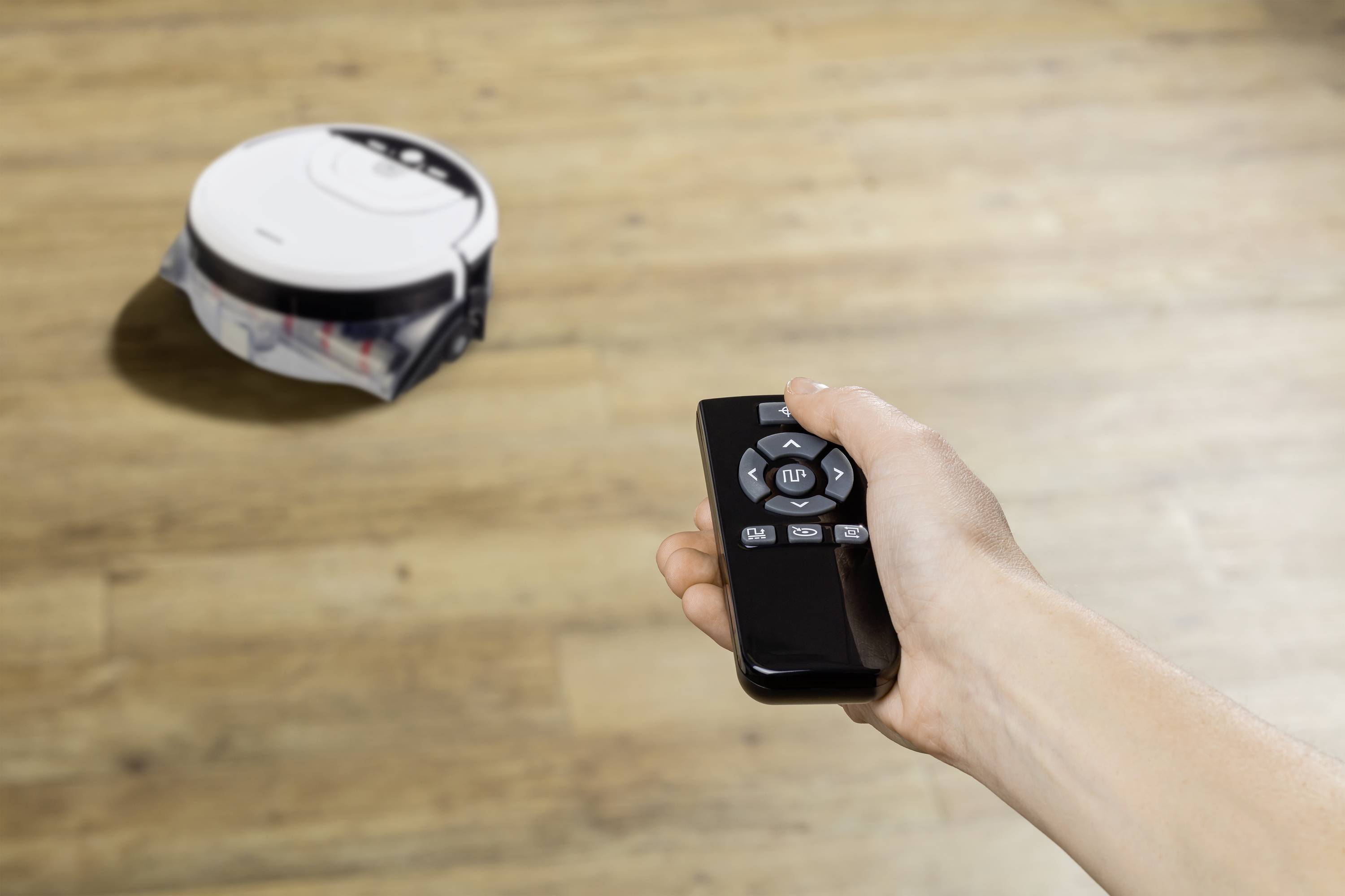 A hand is holding a remote control pointing at a round, white robotic vacuum cleaner moving across a wooden floor.