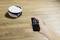 A hand is holding a remote control pointing at a round, white robotic vacuum cleaner moving across a wooden floor.